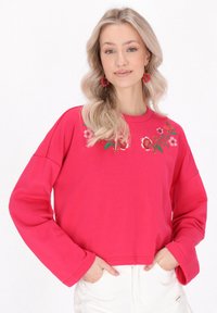 Rózsaszín, rövidített pulóver hosszú ujjakkal, piros és zöld virágmintás hímzéssel. Puha anyag, laza szabás és kerek nyakkivágás.