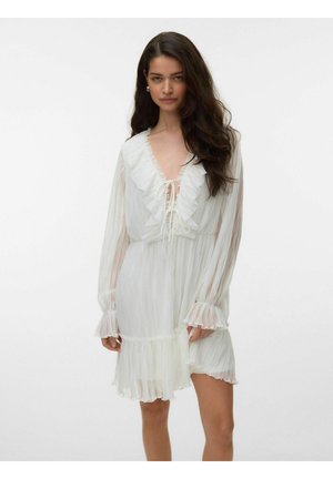 Femme aux longs cheveux foncés portant une robe blanche plissée avec un col à volants, un laçage à l'avant, des manches longues et une jupe courte évasée.
