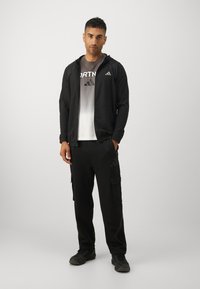 Zwarte sportjack met capuchon, met subtiele textuurdetails. Gecombineerd met zwarte cargobroeken en een grijze t-shirt met een verloop en logo.