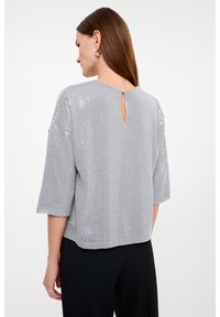 Maglietta a maglia grigia chiaro e testurizzata con maniche corte, caratterizzata da un dettaglio a chiave sulla schiena e piccole paillettes per un tocco di brillantezza. Abbinata a pantaloni neri.