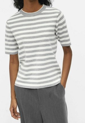 Personne portant un haut en maille à manches courtes rayé blanc et gris avec un pantalon gris sur mesure, main glissée dans la poche, sur un fond uni.