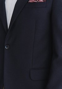 Granatowa marynarka o fakturze materiału, z pojedynczym zapięciem na guzik, boczną kieszenią oraz różową, wzorzystą poszetką w kieszeni na piersiach.