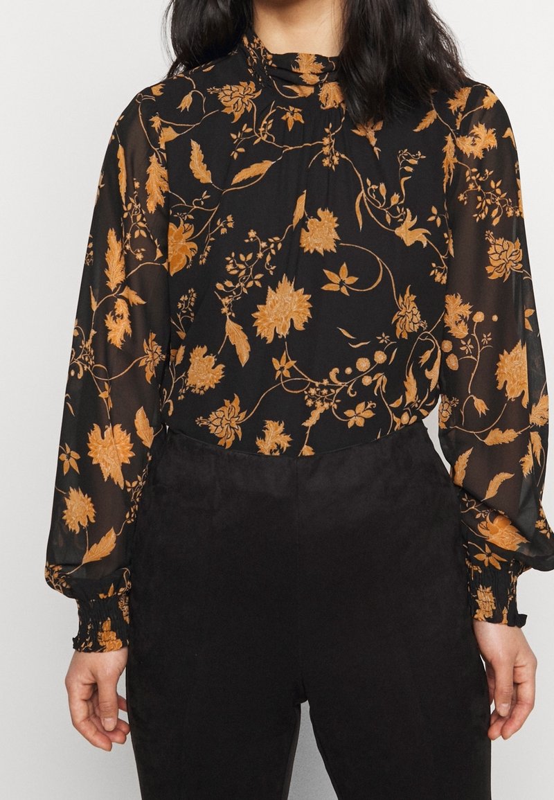 Femme portant un chemisier noir transparent à manches longues avec des motifs floraux orange, associé à un pantalon noir taille haute.