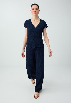 Femme marchant vers l’avant portant un haut cache-cœur texturé bleu marine et un pantalon à jambes larges avec des sandales beige à bout ouvert sur un fond clair uni.