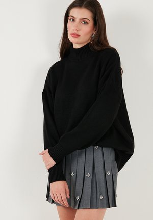 Zwarte gebreide trui met een hoge hals en verlaagde schouders, gecombineerd met een grijze geplooide rok met decoratieve bloemversieringen.