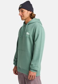 Felpa verde in cotone con tasca a marsupio, cappuccio con cordino regolabile e grafica stampata con la scritta "CALMATI, SII ACQUA." Indossata con un berretto navy.