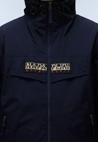 Granatowa kurtka na zamek z teksturowanego materiału; posiada dwie górne kieszenie z logo "NAPAPIJRI" oraz regulowany kaptur ze ściągaczem.