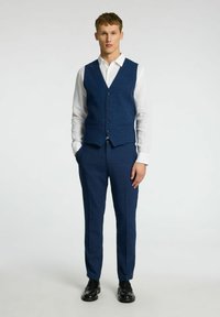 Temno modra brezrokavnik in usklajene hlače iz teksturirane tkanine. Bela srajca spodaj, črne čevlje in podrobnost s šestimi gumbi.