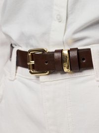 Cinto de couro castanho com uma fivela dourada polida, de formato retangular e textura suave, usado sobre calças brancas de ganga.