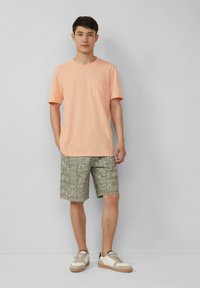 Licht koraalkleurig T-shirt met korte mouwen en een borstzak, gecombineerd met geprinte olijfgroene shorts en beige sneakers met witte accenten.