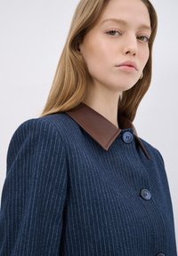 Námořnický pruhovaný kabát s hnědým koženým lemováním, se dvěma modrými knoflíky a přiléhavým střihem, vyrobený z texturované tkaniny.