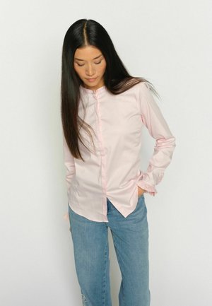 Ženska z dolgimi temnimi lasmi, oblečena v svetlo rožnato srajco z gumbi in modre kavbojke, stoji z eno roko v žepu, zre navzdol.