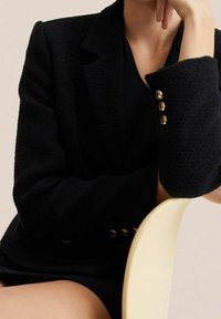 Czarna teksturowana marynarka z klapami i trzema złotymi guzikami na rękawach. Nosi się ją na ciemnej bluzce, w połączeniu z prostym krzesłem.
