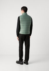 Vest verde din fâșie cu cusături orizontale, guler înalt, purtat peste o cămașă neagră cu mânecă lungă, asortat cu pantaloni negri și pantofi masivi.