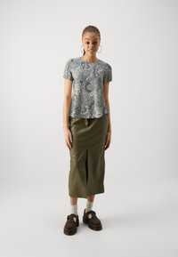 Bluză cu mâneci scurte, de culoare teal, cu modele paisley, asortată cu o fustă de lungime medie, de culoare verde măsliniu, cu o despicătură frontală, purtată cu pantofi maro și șosete gri.