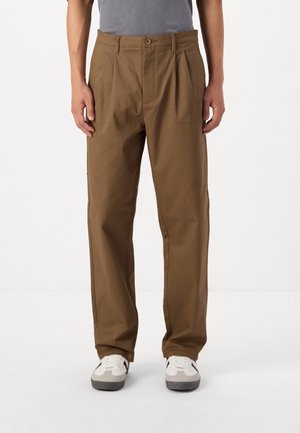 Brown, pleated trousers made from a sturdy fabric, featuring a button closure, side pockets, and a relaxed fit, paired with white sneakers.