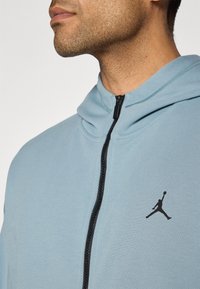 Lyseblå zip-up hoodie med høj krave, sort lynlås og sort Jordan-logo på venstre brystområde. Blødt stof.