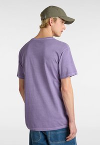 T-shirt di cotone viola con maniche corte e scollatura a girocollo, abbinata a pantaloni corti di jeans blu. Viene indossato un cappellino verde, dotato di cinturino regolabile.