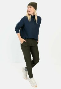 Marine gebreide trui, olijfgroene broek en beige sneakers. Het model poseert met de handen in de zakken, met een geribde beanie en een ontspannen pasvorm.