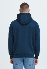 Navyblå hettegenser med snorhette, lange ermer og ribbestrikkede mansjetter. Har en glatt tekstur og en avslappet passform, kombinert med lyse denimjeans.
