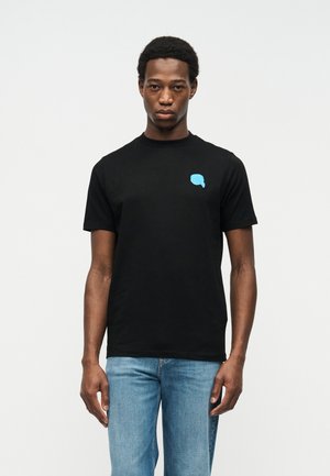 Homme portant un t-shirt noir avec un petit graphique de cerveau bleu sur la poitrine, associé à un jean bleu clair, debout devant un fond blanc uni.