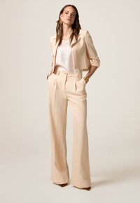 Hellbeige, verkürzter Blazer mit hochgeschlagenen Ärmeln, kombiniert mit hochtaillierten, plissierten Hosen. Das Modell trägt ein weißes Satin-Oberteil und gemusterte Absätze.