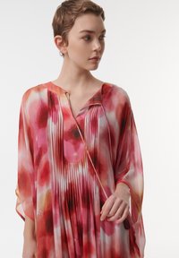 Transparante, vloeiende kaftan in levendige rood- en tinten roze met een aquarelpatroon. Heeft plooien en een ronde halslijn, met vleugelachtige mouwen.