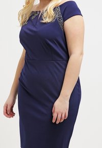 Námořnická modrá úzká šaty s krátkými rukávy bez ramínek, zdobené aplikacemi na ramenou a hladkým povrchem.