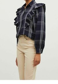 Navy plaid blouse with ruffled front and shoulders, long sleeves with cuff, made of lightweight fabric, paired with fitted beige pants.