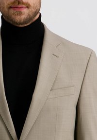 Helle beigefarbene Sakkos mit strukturiertem Finish und schlanker Passform, gepaart mit einem schwarzen Rollkragenpullover darunter. Sichtbare Brusttasche und Reversdetails.