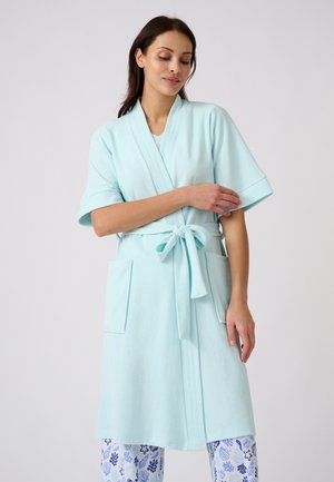 Vrouw draagt een lichtblauwe kamerjas met korte mouwen, gestrikt in de taille met grote zakken, gecombineerd met witte en blauwe broek met patroon.