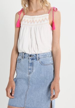 Kvinde iført hvid ærmeløs top med lyserøde kvastbindinger og broderi, kombineret med lysblåt denimskørte med frontslids.