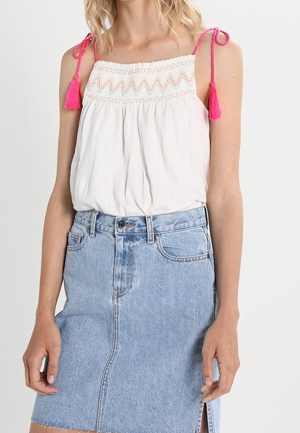 Femme portant un haut sans manches blanc avec des liens à pompons roses et une broderie, associé à une jupe en jean bleu clair avec une fente à l'avant.