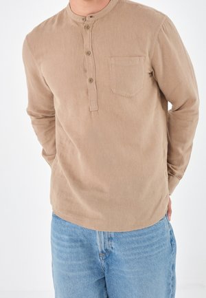 Homme portant un henley beige à manches longues avec des boutons et une poche poitrine, associé à un jean denim bleu clair, les mains derrière le dos.