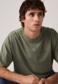 Νέος άνδρας με καστανά σγουρά μαλλιά, φορώντας ένα χακί πράσινο T-shirt Levi’s, καθισμένος με τα χέρια σταυρωμένα και κοιτάζει ελαφρώς προς τα πλάγια.