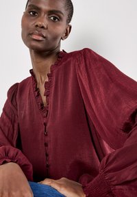 Donna con capelli corti che indossa una blusa bordeaux con volant, bottoni e maniche a sbuffo, appoggiando una mano sul ginocchio.