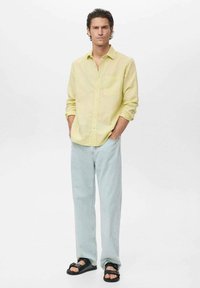 Camisa amarilla de lino de botones, jeans de mezclilla azul claro y sandalias negras. La camisa tiene un corte relajado y las mangas enrolladas.