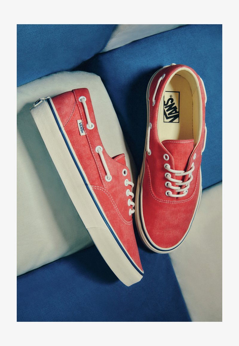 Baskets Vans rouges avec lacets et semelles blanches, posées sur des coussins bleus et beiges. Une chaussure est à plat; l’autre est posée sur le côté.