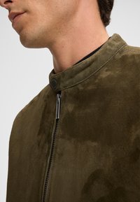 Olijfgroene suède jas met een ronde kraag, rits aan de voorkant en drukknoop. Glad oppervlak met een subtiele glans en zonder zichtbare patronen.