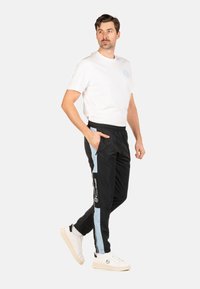 Homme portant un t-shirt blanc et un pantalon de jogging noir avec des rayures latérales bleu clair et des baskets blanches, se tenant avec une main dans sa poche.