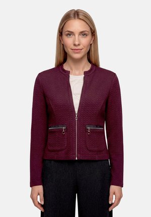 Femme portant une veste zippée texturée violet foncé avec des poches zippées, au-dessus d'un haut blanc et d'un pantalon noir, debout devant un fond uni.