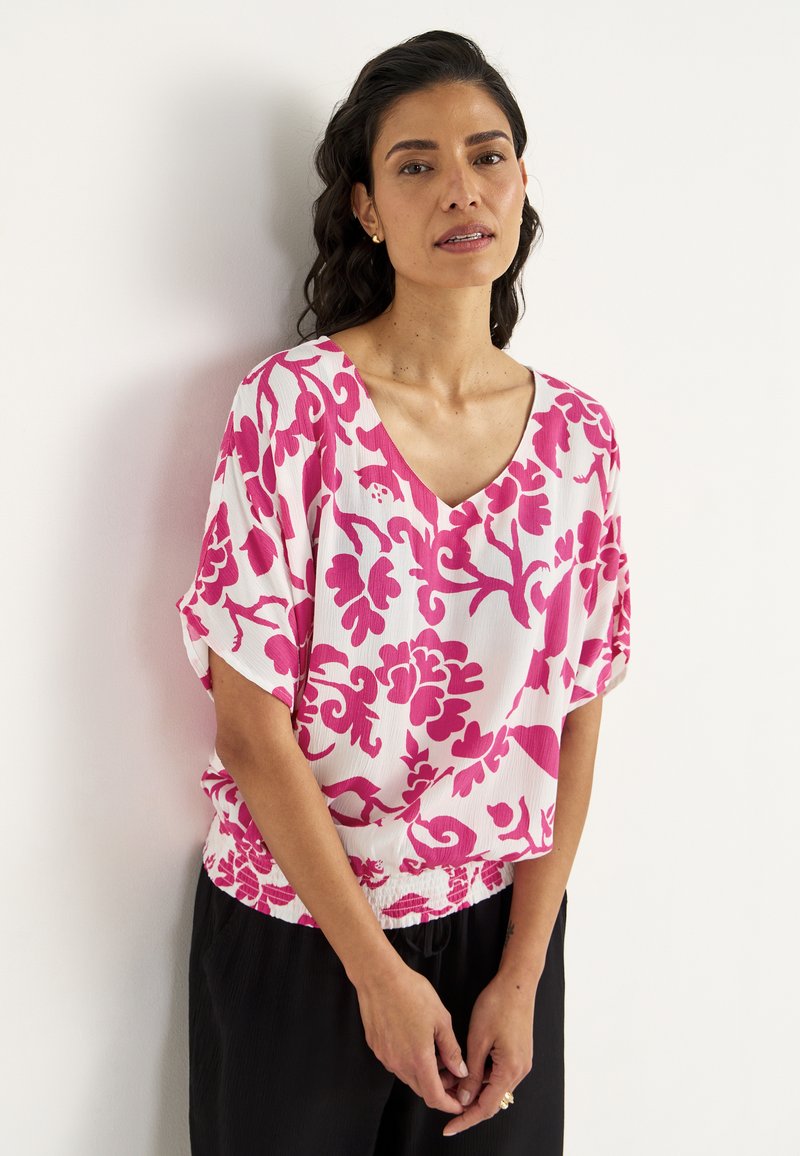 Korte mouwen blouse in het wit met een gedurfde roze bloemenpatroon, v-hals ontwerp en een geraffineerde taille, gecombineerd met zwarte broeken.