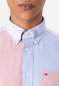 Camicia con colletto con pannelli verticali a strisce blu e rosa. Presenta una chiusura con bottoni e una piccola tasca con logo. Tessuto con texture.