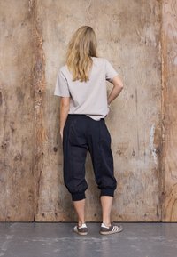 Vrouw met lang blond haar, gekleed in een beige T-shirt, zwarte cropped broek en bruine sneakers, staat met één hand op haar heup tegenover een houten muur.