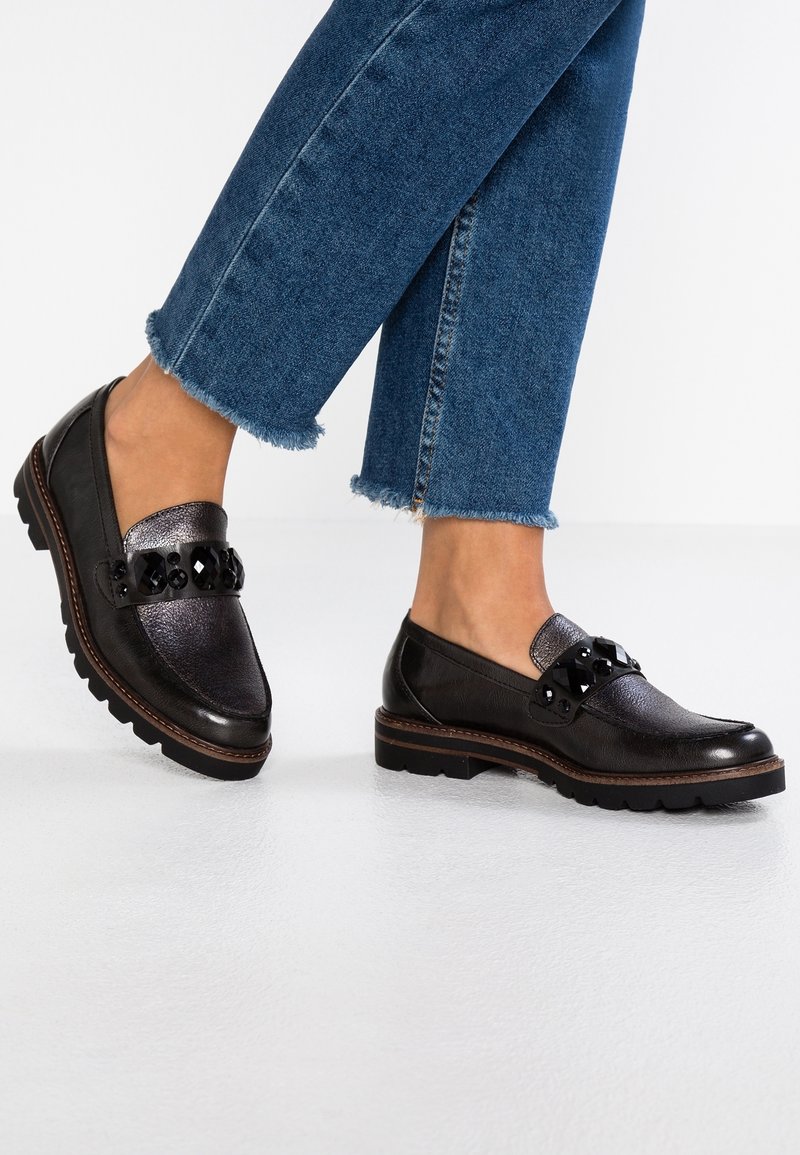 Mocassins en cuir noir avec semelles épaisses et grosses décorations en pierres noires, portés avec un jean bleu à ourlet effiloché.