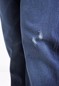 Jeans in denim blu scuro con ginocchio consumato e due piccoli fori sfrangiati. Il tessuto ha una texture liscia e un design classico con cinque tasche.