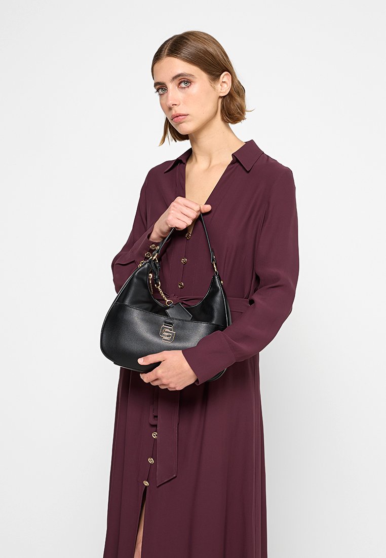 Borsa a tracolla in pelle nera con forma curvata, dettaglio del logo dorato e catena decorativa sulla tracolla. Il modello indossa un lungo vestito bordeaux.