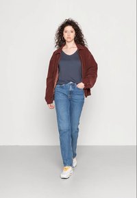 Rjava kordurojska jakna, mornarsko modra majica, modri kavbojke in bele superge z rumenimi detajli. Model stoji ob običajni podlagi.