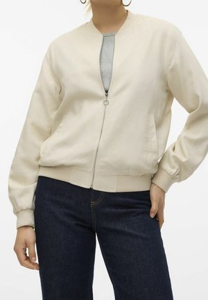 Persona que lleva una chaqueta bomber cremosa con cremallera, camisa gris, vaqueros azul oscuro y pendientes de aro dorados, posando con una mano en la cadera.