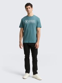 Teal katoenen t-shirt met korte mouwen en witte printdetails, gecombineerd met zwarte jeans en witte sneakers. Eenvoudig, casual ontwerp.